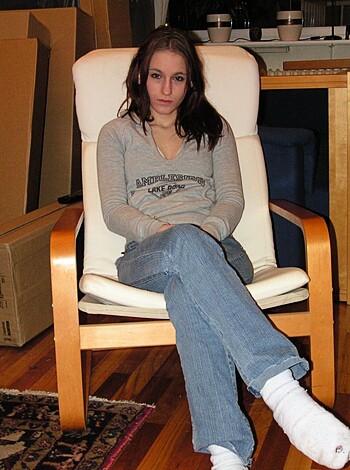 A blonde chick is perched on her ass cheeks, straddling a plush armchair while flaunting her skinny legs in tight blue denim shorts with a low-cut top revealing ample bosom.