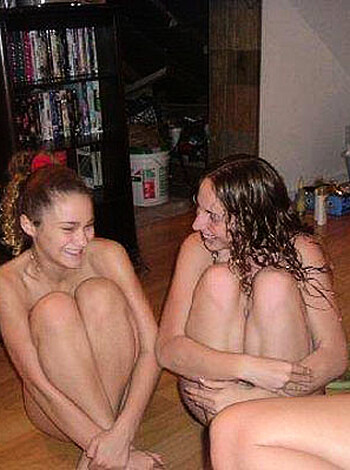 Two completely nude chicks laughing their asses off while sitting on the couch in someone's living room.