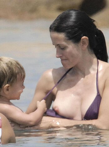 An attractive bare-boobed babe holding her baby's tiny fist while gazing downward with a delightful smile on her lips at the sandy beach.