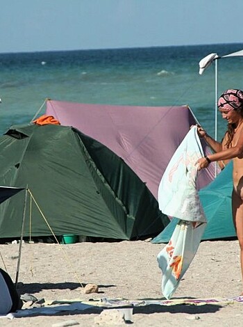 A bikini-clad babe wiping her pussy while some dude's got his back to the camera. Beach dudes chillin' in the background.