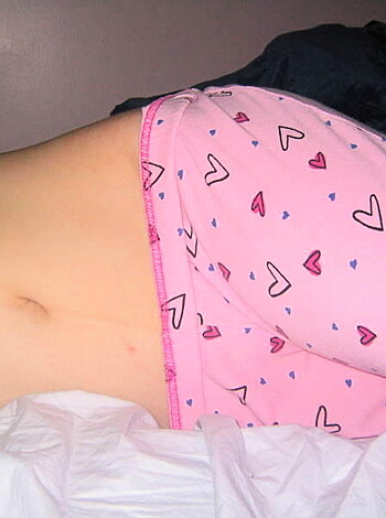 Image of a chick with her cunt hairs sticking out, lying down on a bed with a giant pink fucking heart-shaped thong on, spreading her legs wide open and showing off her bald snatch.