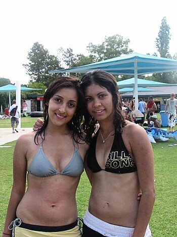 Here's the description of the image: "Two hot chicks taking a selfie in the pool area. One with tits hanging out and a skimpy bikini on, the other showing off her slender legs in a cheeky bathing suit."