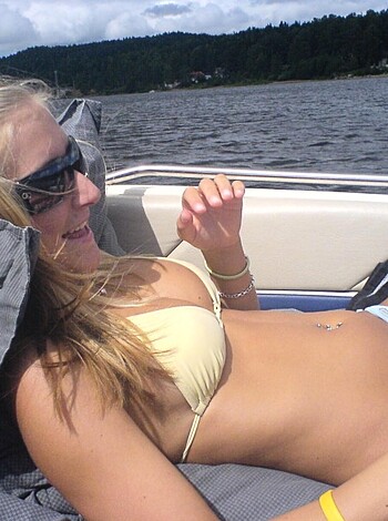 A fuckin' hot blonde chick in a tight-ass yellow bikini top and matching blue bottoms is lying back on the ass-end of a boat, grinnin' like a fuckin' idiot while gettin' some sweet sunbathing action with her legs crossed.