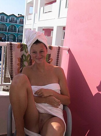 Here's a hot vulgar shot of a topless chick sitting on a chair, her tits out and her pussy exposed, holding a towel over her crotch for modesty while she grins at the lens.