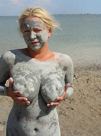 Image depicting naked woman lying on sandy shore with face upwards, covered in muddy waters.