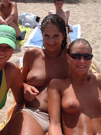 Bare-breasted hotties grinning for the camera while sitting on a beach in their skimpy swimwear.