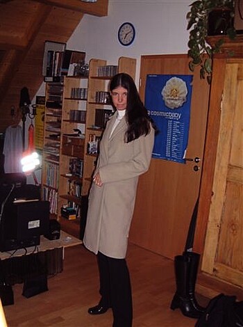 A fucking stressed-out bitch stands outside her goddamn book-filled living space, wearing a motherfucking trench coat, while gazing at the camera with a look of complete uncertainty or discomfort on her face.
