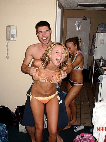 A chick in a bikini's tits are out 'cause two dudes are makin' funny faces in front of their pals, in a living room.