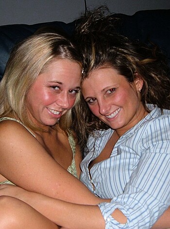 Two slutty broads chillin' on a couch, one with her tits out for all to see, grinnin' like fools.