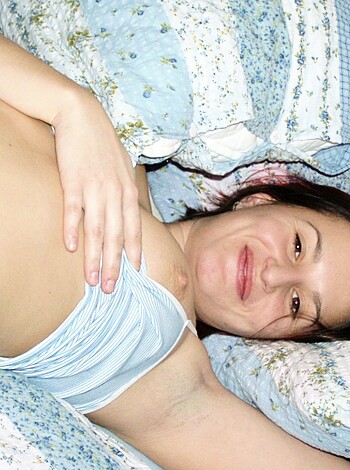 The image shows a hot naked babe relaxing on a bed, flashing her tits for the camera with a big smile on her face.