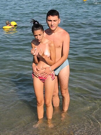 Two dudes standin' nekkid on the shoreline of a beach, balls hangin' out like chicken wings.
