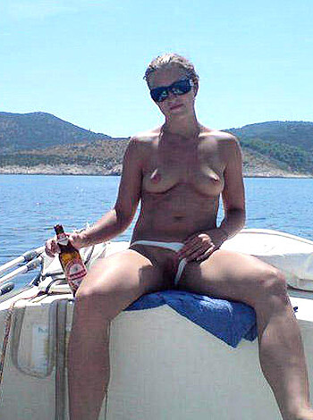 Here's the vulgar, factual description of the image without any introduction or greeting or other: 
"The photo shows a busty blonde chick with ample tits and big ass sitting on a boat in just a skimpy yellow bikini bottom that barely covers her pussy. She's holding a cold beer, her hair is messy, and she looks drunk as fuck."