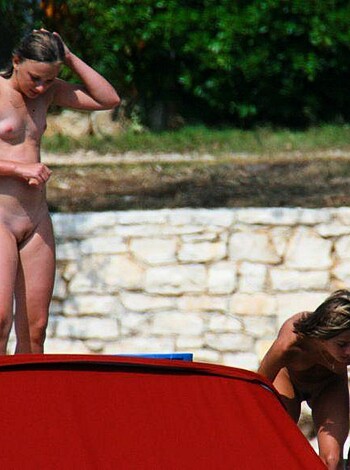 "Two tits-out chicks sunbathe on the sand, the standin' one showin' off her rack while sittin', bustin' out her boobs."