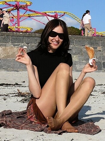 Here's the vulgar description of the image: A busty bitch is sitting on a sandy beach, sporting a tight black dress and shades, while munching down on a fuckin' ice cream cone like a goddamn pro.