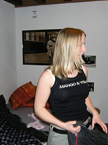 Here's a vulgar description of the image: "Chick in a sports bra and camo pants, bending over next to some fucking bed with white walls and shitty posters on them."