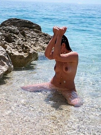 A fuckin hot chick is sittin' ass-naked on some rocks by the beach, her tits bobbin' up and down with the waves like a goddamn porno movie.
