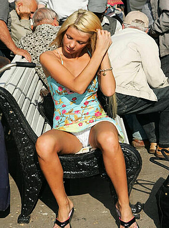 The blonde lady is posing seductively while resting her hand on her floral dress covered thigh and revealing her sexy thigh high stockings and shoes as she sits on a black bench.