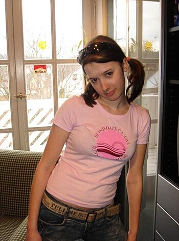 The image depicts a blonde bitch, dressed in a pink tee and denim shorts, striking a pose while grinning her fake-ass smile to the camera.