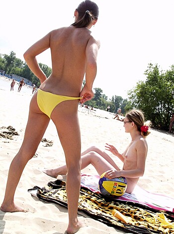 Two chicks at the beach, one sittin' ass-down while the other stands tall in her yellow bikini bottoms, showing off those hot legs of hers!
