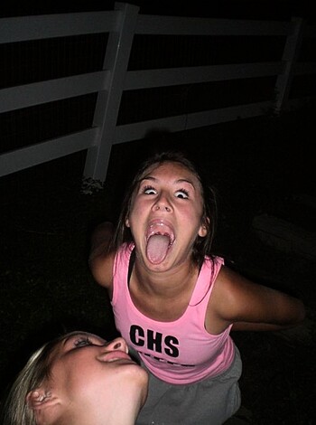 This fuckin' picture shows two hot, young chicks in an outdoor setting at night, one of 'em's got her tongue out licking her lips or like maybe bitin' some pussy above her head, while the other one's standin' beside her with a blank expression. Shit looks like they're havin' fun and fuckin' around playful-like.