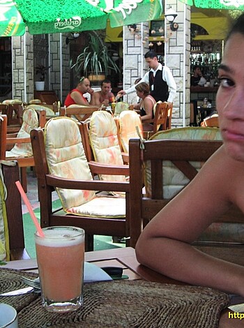 A bikini-clad broad sits at an outdoor cafe, elbow resting on a tray while her chin's on it, deep in thought or jerking off while some guys eat their meals nearby and a waiter tries to get her attention.