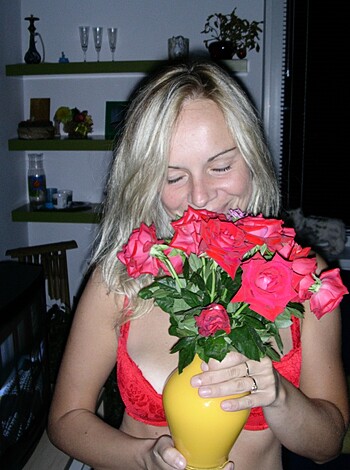 Vulgar description: "Look at this fucking hot babe in a sexy red bra with tits out, showing off her shiny pink lips while holding some flowers up to her face like a goddamn model."