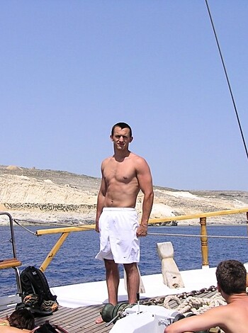A half-naked dude, probably on vacation, is posing for the camera while standing shirtless on a speedboat. He's got some serious tan lines going on and seems to be quite proud of his chiseled abs.