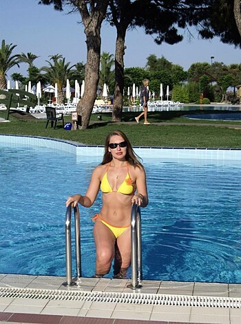 "A busty brunette with a stripper pole sits on the edge of a glittering blue pool, flashing her ample titties while showing off her tanned pussy."