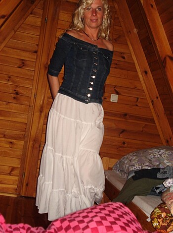 Here's a description of the image of some bitch in a fuckin' denim skirt and a prissy white blouse, standing inside a shitty little cabin-like room. 30 words.