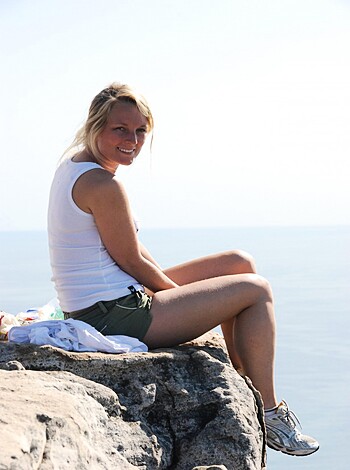 Here's a vulgar description of this image: 
A busty blonde in a white tank top and daisy duke shorts is sittin' on her ass cheeks on a fuckin' rocky cliff overlookin' the goddamn ocean, with a cockblockin' view of that sweet pussy.