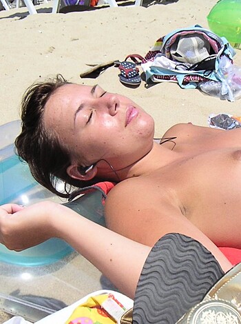 Image of a busty blonde laying on a beach towel, boobs out, listening to music while chilling at the beach with her hand resting on her head.