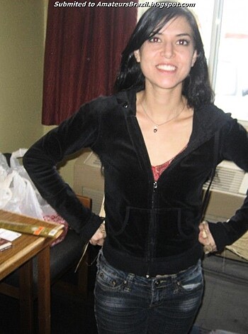 A nude woman is standing in front of a camera with her legs spread apart and her hand placed on the wall, all while wearing a black jacket that is zipped up to mid-thigh, exposing her shaven pussy and clit.