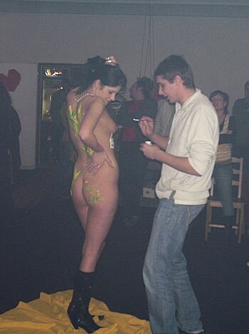 A naked bitch is covered in colorful shit while some dickhead reads a book with his hand up her fucking ass, gesturing towards the audience.