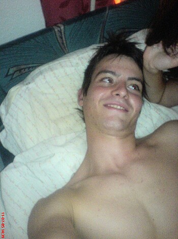 Here's the vulgar description of the image: "A fucking hot dude with a big dick is laying naked on a bed, smiling and showing off his perfect tits, inviting you to suck them off."