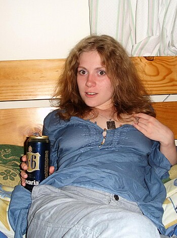 "Young chick lying down on mattress, sipping from brown bottle."