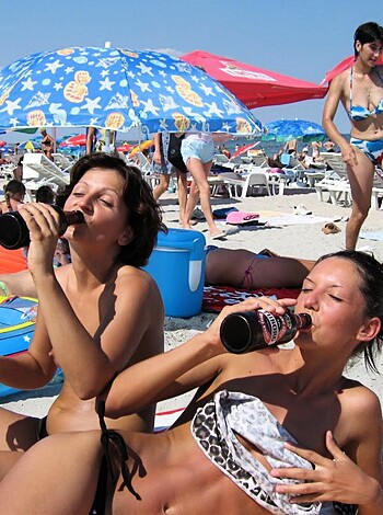 This image depicts two chicks suckin' down some fucking soda while baskin' in the sun with their titties out, all whilst lying on a beach towel surrounded by a bunch of fuckin' umbrellas.