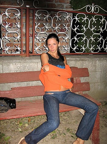 A naked broad is taking a piss on the curb next to some metal benches, wearing nothing but her fuck-me-heels and a tight orange t-shirt with blue jeans. She's got long brown hair flowing down her back and is posing for some dude's camera, smiling seductively while holding onto the top of the bench.