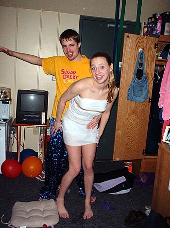 This image is a fucking hilarious scene of a tits-out babe clad in a goddamn toga writhing her ass off while getting spooned by some lazy shit in pink pajamas, both of them lookin' like they're havin' the time of their lives.