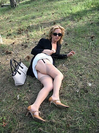 A nude female is displayed with her genitals exposed, wearing high heels and sunglasses while lying down on the ground with her legs splayed apart in a rural area.