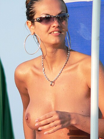 A shapely babe with massive boobies is standin' on the sandy shore, while her titties are bustin' outta her bra, wearing sunglasses and a necklace. There's an umbrella in the background 'cause it's sunny as fuck here.