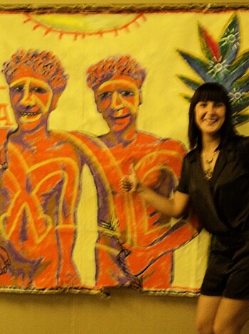 A hot chick stands by a dick pic of two hairy dudes with tats and skulls, in an art exhibit full of fucking nonsense.