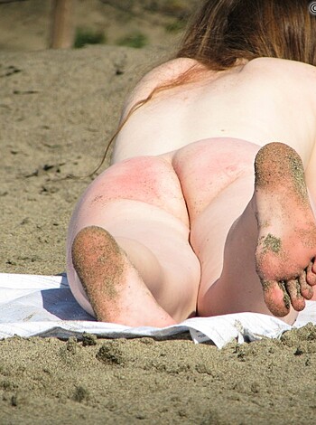 This image depicts an unidentified bikini-clad woman with her hands covering her chest area while she sits on a bright yellow beach towel, completely nude save for the streak of red paint adorning her backside.