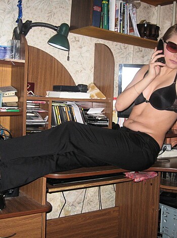 Here's the vulgar description of the image: A slutty bitch with big tits and a bald pussy is lying naked on some dude's office desk, using his fucking phone to talk shit while he probably jerks off in the background.
