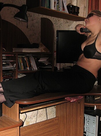 A nude woman sits on a desk, dressed only in black lacy panties and heels.