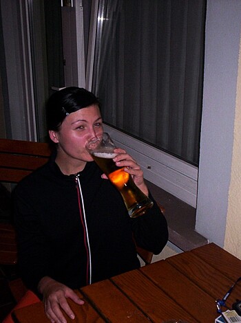 Image depicts busty blonde with massive tits chugging down brews while sitting at the kitchen table, her lips wrapped around the rim of a frosty mug while she tilts back to take in a gulp.