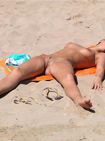 A nude woman lies on a towel, her nipples hard and perky, her legs spread wide open, while basking in the warm sun on a sandy beach.