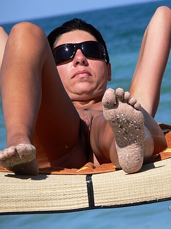 The vulgar description of this image is of a hot chick with an awesome body lying on a lounge chair by the seashore in her skimpy bikini bottoms, showing off her milky white feet and golden beach glow.