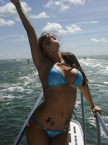 Here's the vulgar description of the image: "A naked chick on a yacht, spreadin' her tits out and laughin', showin' off her snatch."