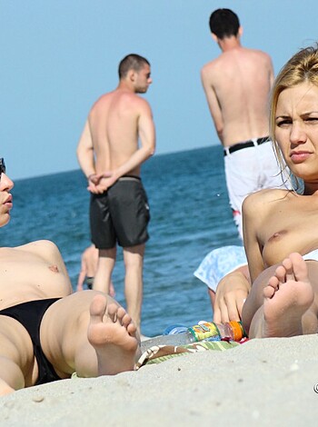 Two naked chicks sunbathing on the sandy shore, with their racks fully exposed for all to see.