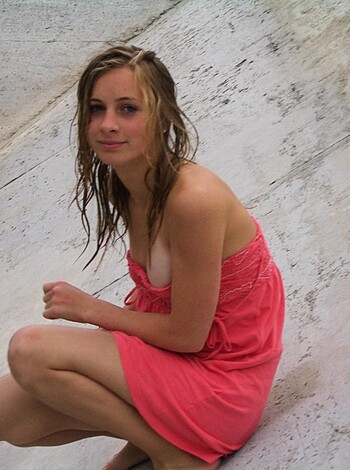 A hot, busty babe in a flirty pink dress is sitting on some rocks with her legs spread and flashing you a toothy grin.